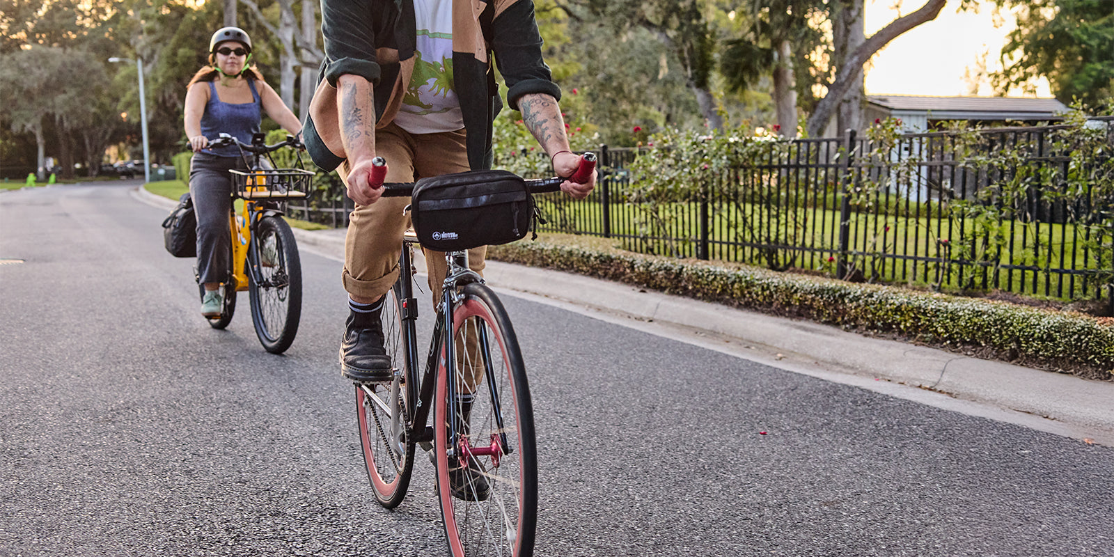 Two people riding bicycles on a paved road with trees and a fence in the background.