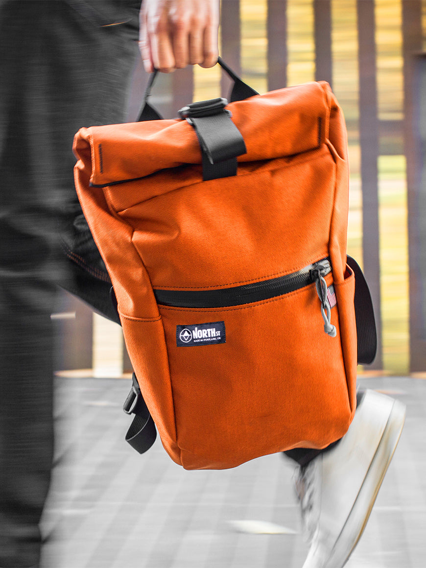 Orange backpack with a visible brand logo held by a person on a blurred  background all-groups